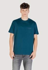 T-shirt in cotone color teal con collo a giro e maniche corte. Presenta un piccolo logo stampato grigio sul lato sinistro del petto. Abbinato a jeans grigi.