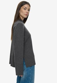 Sivijo pleteni pulover z visokim ovratnikom in rebrastimi manšetami, z ohlapnim krojem in stranskimi rezi, v kombinaciji z svetlo modrimi kavbojkami.