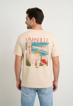 Hombre vistiendo una camiseta beige con una ilustración colorida del paisaje urbano de Marsella, que incluye iglesia, puerto y árboles, combinado con vaqueros azules.