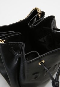 Černá kožená kabela ve tvaru vědra s texturovaným povrchem, zlatými řetízkovými detaily a stahovací šňůrkou. Interiér obsahuje několik přihrádek.