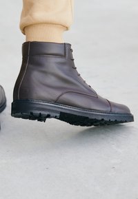 Braune Leder-Ankle-Boots mit sichtbaren Schnürsenkeln, robuster Sohle und strukturiertem schwarzen Gummiaußensohle, die ein schlankes, minimalistisches Design aufweisen.
