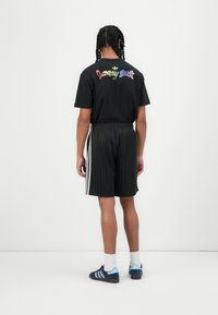 Black cotton t-shirt with multicolored "Jeremy Scott" text on the back; paired with black pinstriped shorts and blue sneakers with white accents.