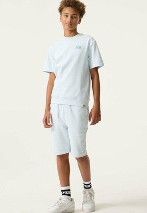 Niño con cabello rizado que lleva pantalones cortos azul pálido y sudadera de manga corta a juego, zapatillas blancas y calcetines blanco y negro, de pie sobre fondo blanco.