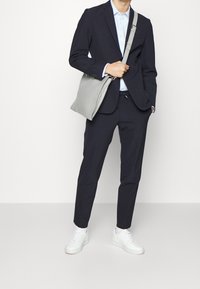 Marinblå kostym med skräddarsydd passform, tillsammans med en ljusblå skjorta och vita sneakers. En grå axelväska hålls i vänster hand.