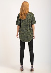 Tarnfarbenes Hemd mit kurzen Ärmeln, abgerundetem Saum und lockerer Passform, kombiniert mit schwarzen engen Hosen und grauen High Heels, von hinten betrachtet.