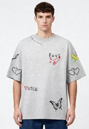 Giovane uomo con una T-shirt grigia oversize con disegni grafici neri e rossi di cuori, una farfalla, un cerotto e parole come "LOVE" e "COUTCH".