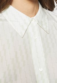 Chemise vert clair semi-transparente présentant un motif en zigzag, un col pointu et des boutons blancs, avec une finition en tissu texturé.