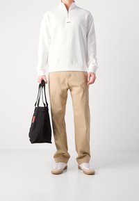Felpa bianca con zip e logo, pantaloni beige e borsa tote nera con dettagli rossi. Sneakers bianche completano l'outfit.