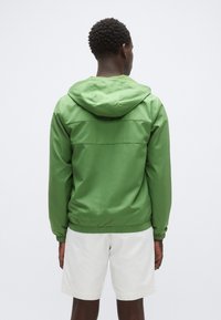 Zelena jakna s kapuco z gladko teksturo, elastičnimi manšetami in vrvico. Na zgornjem delu hrbta ima vidno šivano obliko.