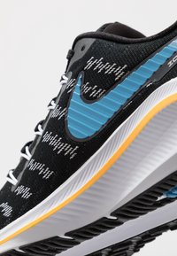 Ingrandimento di una sneaker nera con il logo Nike blu, caratterizzata da un tessuto testurizzato e una suola bianca con un'accents arancione.