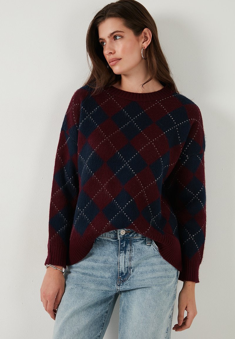 Pull en maille bordeaux et marine avec des motifs en diamants, encolure ronde, coupe ample, ourlet côtelé et manches longues. Associé à un jean bleu clair.