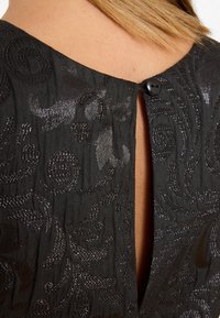 Gros plan d'une robe en tissu texturé noir avec des motifs tourbillonnants, fermée par un seul bouton noir au niveau de l'encolure arrière au-dessus d'une fente ouverte.
