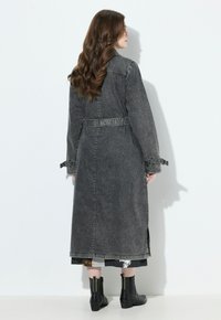 Žena s dlouhými vlnitými vlasy, oblečená v černém opraném džínovém dlouhém kabátu a černých kotníkových botách, stojící bokem proti bílé zdi.