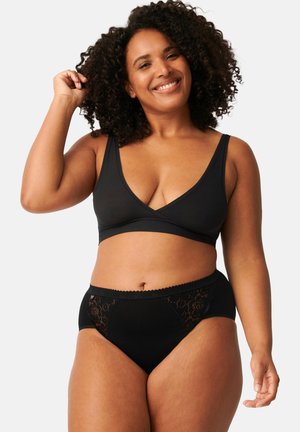 Donna sorridente con capelli ricci che indossa un bralette nero e mutandine nere con dettagli in pizzo su uno sfondo semplice.