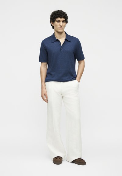 Άνδρας που φοράει μπλε σκούρο πόλο με κοντά μανίκια, λευκό παντελόνι φαρδύ και καφέ παπούτσια, στέκεται με το ένα χέρι στην τσέπη.