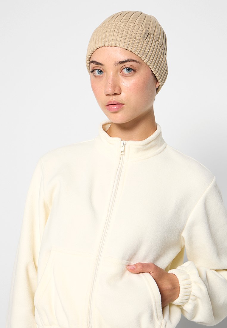 Beżowa dzianinowa czapka z prążkami noszona z białą polarową kurtką na zamek. Czapka ma subtelne pionowe linie i dopasowany krój.