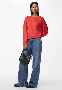 Sudadera cortada roja con mangas amplias, combinada con unos jeans de mezclilla sueltos y de color azul claro y un pequeño bolso negro. Calzado negro completa el look.