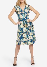 Kvinna i knälång blå blommig omslagsklänning med volangärmar och fåll, knuten midja, bär svart ankelsandaler med remmar, händerna på höfterna.