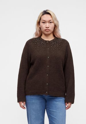 Person står med fronten mod kameraet iført en mørkebrun knappet cardigan med små perledetaljer og lyseblå jeans mod en ensfarvet baggrund.