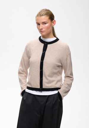 Femme aux cheveux blonds en queue de cheval porte un cardigan beige avec bordure noire, boutonné par-dessus une chemise blanche et un pantalon noir, les mains dans les poches.