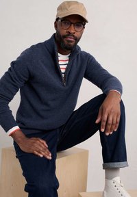 Mörkblå zippered tröja med texturerad stickning, röd och vit randig skjorta under, mörka jeans, beige keps och vita högstövlar.