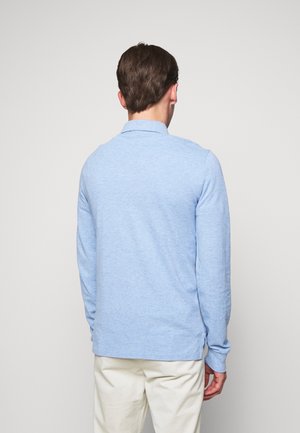 Mann trägt ein hellblaues langärmeliges Poloshirt und weiße Hose, steht mit dem Rücken zur Kamera vor einem schlichten hellen Hintergrund.