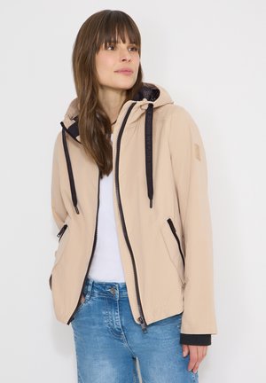 Frau mit glattem braunem Haar, die eine beigefarbene Kapuzenjacke mit schwarzen Reißverschlussdetails über einem weißen Hemd und blauer Jeans trägt, blickt nach links.