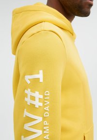 Mann trägt gelben Hoodie mit weißem, erhabenem Text "WT #1" und "CAMP DAVID" auf dem Ärmel, schaut nach rechts.