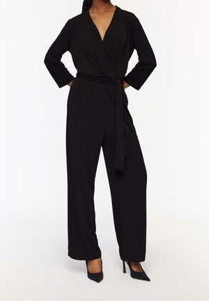 Vrouw draagt een zwarte lange mouwen overslag jumpsuit met riem en zwarte puntige hoge hakken tegen een witte achtergrond.