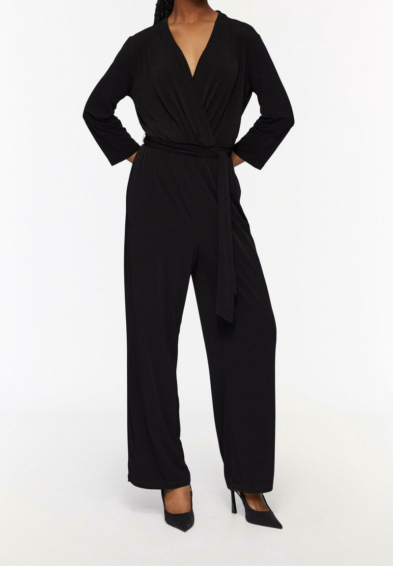 Femme portant une combinaison-pantalon noire à manches longues avec ceinture et des talons hauts pointus noirs sur fond blanc.
