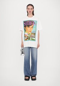 Oversized white t-shirt featuring a colorful magazine cover graphic. Paired with light blue wide-leg jeans and chunky black sandals. Silver handbag.