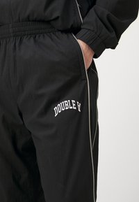 Melni sporta bikses no viegla auduma, aprīkotas ar elastīgu jostasvietu, sānu kabatām un baltu izšuvumu ar "DOUBLE A" logo.