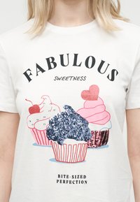 Λευκό μπλουζάκι με τρία διακοσμημένα cupcakes και κείμενο «Υπέροχη Γλυκύτητα Μπουκιά-Μπουκιά Τέλεια» φορεμένο από άτομο με ξανθά μαλλιά.