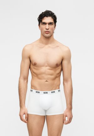 Férfi rövid sötét hajjal, fehér GCDS boxeralsót viselve, egy egyszínű világosszürke háttér előtt áll.