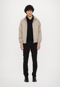 Beige bomberjacka med dragkedja framtill, två sidofickor och ribbade ärmslut, bärs över en svart polo och svarta byxor.