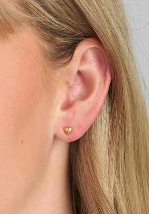 Boucle d'oreille clou en forme de cœur en or avec une surface texturée, positionnée dans une oreille percée, mettant en valeur les contours de l'oreille et la coiffure.