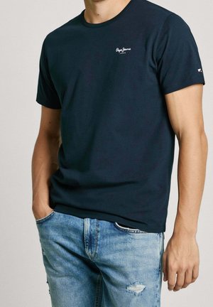 Homme portant un t-shirt uni bleu marine Pepe Jeans et un jean bleu clair déchiré, une main dans la poche, sur fond neutre.