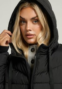 Veste rembourrée noire avec capuche, dotée d'une fermeture éclair et d'un tissu texturé. Portée sur un haut boutonné gris clair.