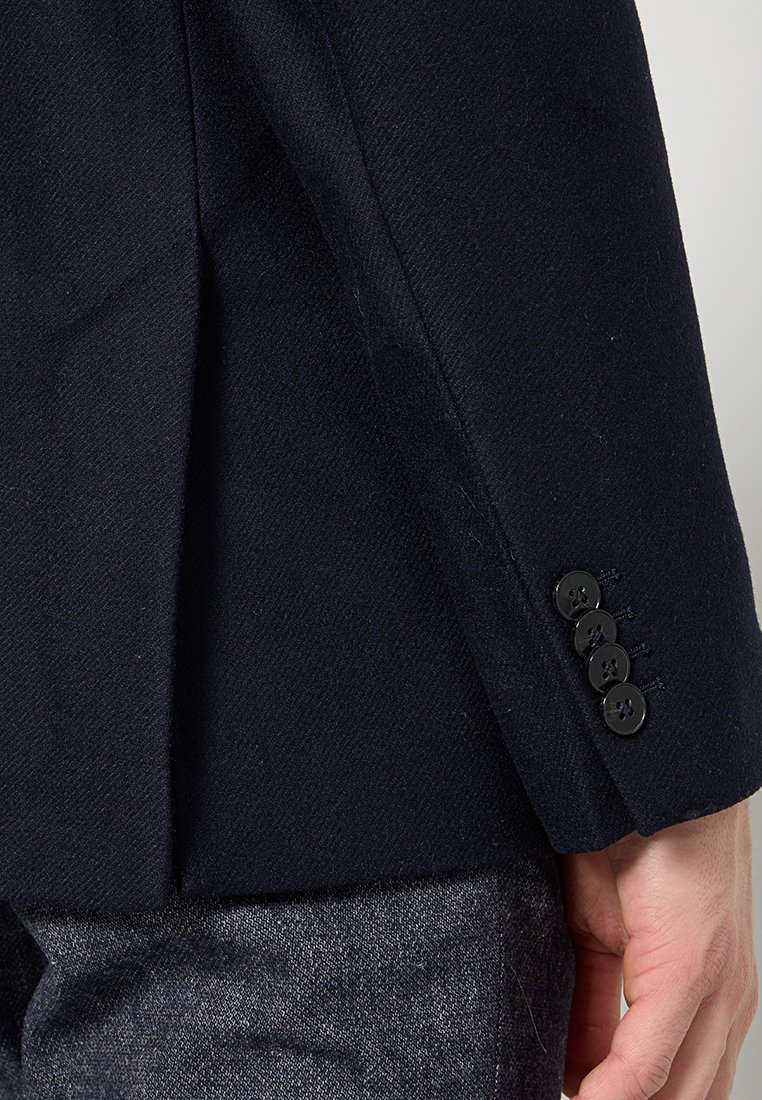 Zbliżenie na rękaw ciemnoniebieskiego żakietu z czterema czarnymi guzikami, położonymi nad ręką spoczywającą obok szarych, fakturowanych spodni.