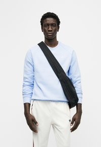 Άνδρας που φοράει ανοιχτό μπλε φούτερ, λευκή φόρμα Adidas με ρίγες και μαύρη τσάντα χιαστί, στέκεται μπροστά σε λευκό φόντο.