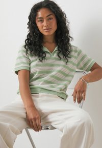 Sieviete ar cirtainiem matiem, tērpusies zaļā un baltā svītrainā īss piedurkņu topā un baltās biksēs, sēž uz caurspīdīgas krēsla pie baltas sienas.