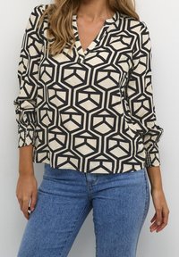 Femme portant un haut à motifs géométriques noir et blanc avec des poignets smockés et un jean bleu, se tenant devant un mur blanc uni.