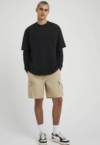 T-shirt noire oversized, short cargo beige avec poches latérales, chaussettes blanches et baskets noir et blanc, debout sur un fond uni.