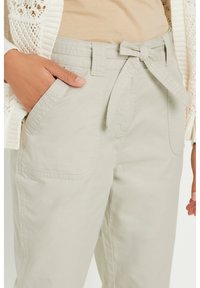 Light beige cotton pants with a waist tie, featuring two side pockets and a smooth texture. A knit cardigan is visible in the background.
