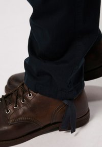 Bruine leren laarzen met veters en metalen oogjes, gecombineerd met donkerblauwe broek met een lichte omslag en blootgestelde stoffen strikjes aan de zoom.