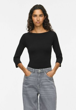 Femme aux cheveux bouclés portant un haut noir ajusté à manches longues et un jean gris taille haute, les mains dans les poches, sur un fond uni.