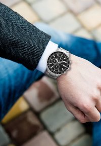 Orologio in metallo argento con quadrante nero, sottodial di cronografo e bracciale in acciaio inossidabile. Il cinturino presenta una finitura spazzolata.