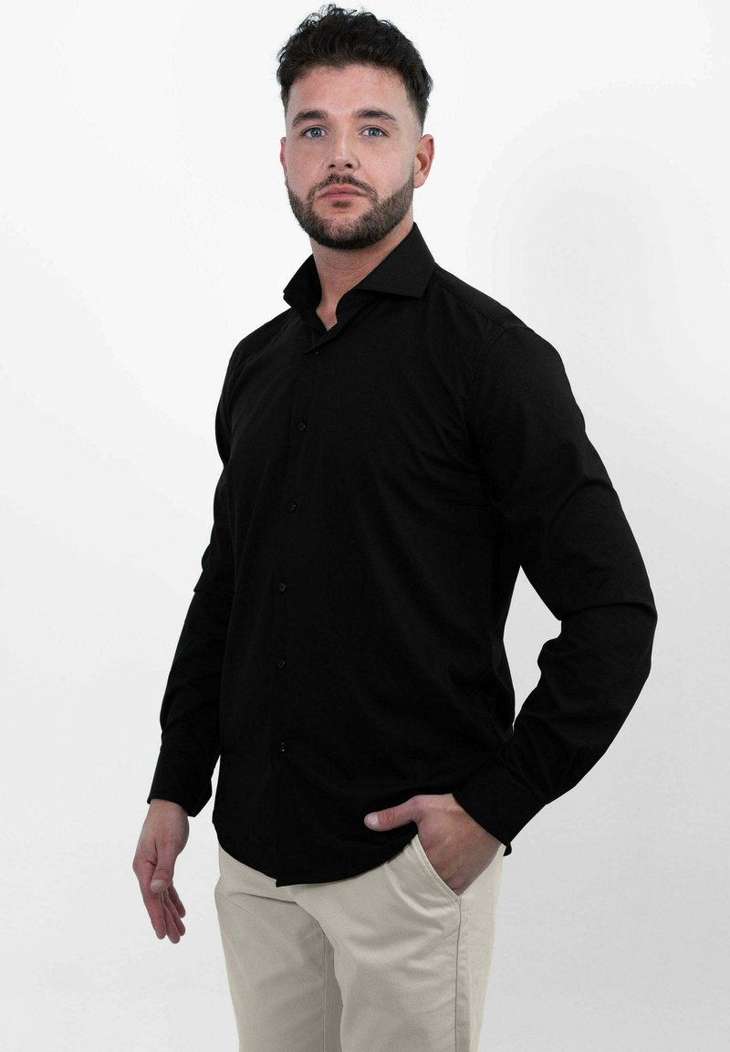 Man met kort donker haar en een baard, gekleed in een zwart knoopshirt en beige broek, staande tegen een witte achtergrond met één hand in zijn zak.