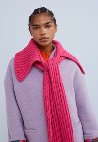 Fular roz tricotat așezat peste o haină de lână lavandă cu guler structurat; țesătură texturată; culori vibrante; mâneci accentuate cu mai multe modele.