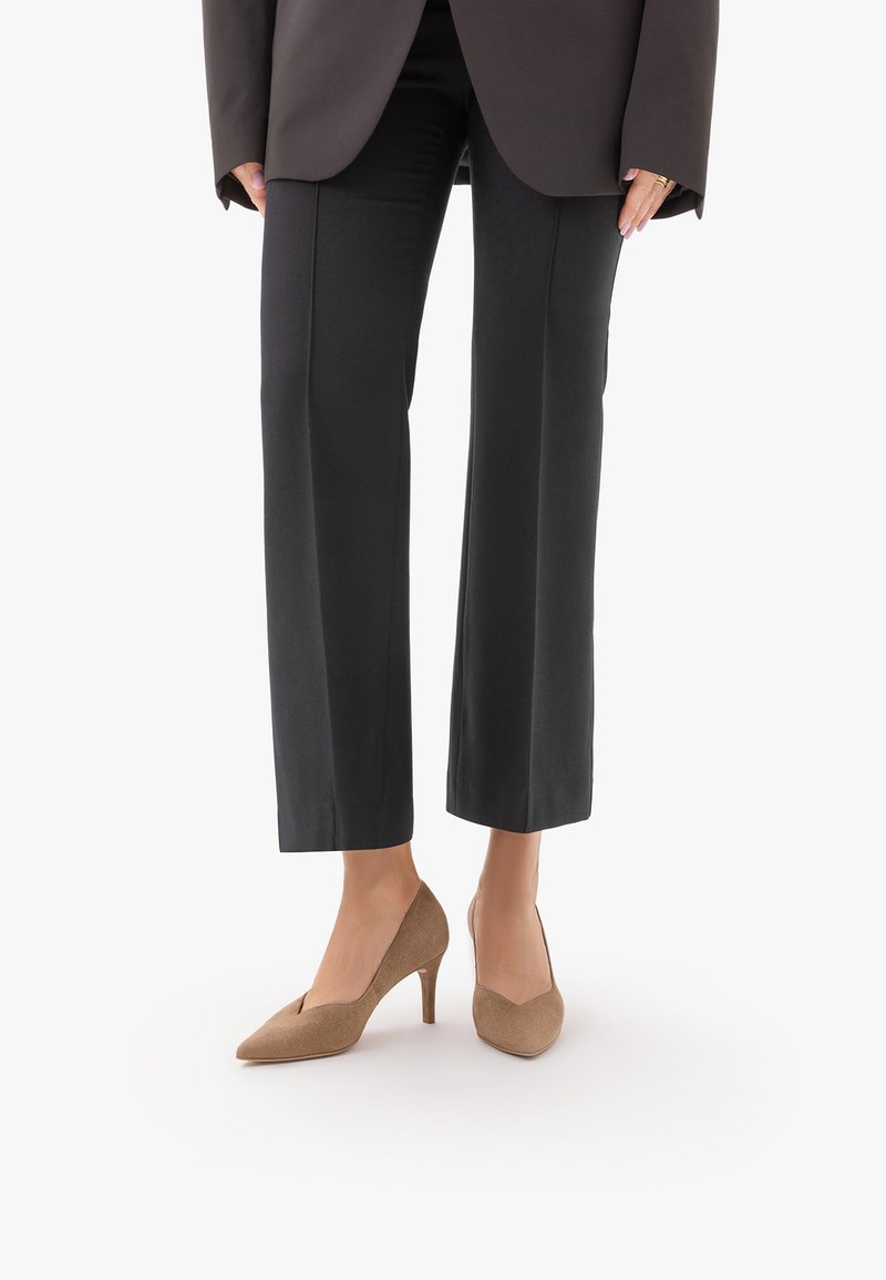 Femme portant un pantalon noir à jambes larges et des talons hauts marron à bouts pointus, debout sur un fond blanc.
